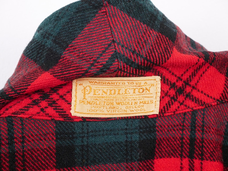 May include: Close-up of a red, black, and green plaid wool fabric. The fabric has a Pendleton label with text that reads "Warranted to be a Pendleton" and "100% Virgin Wool".