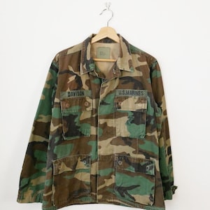 Könnte beinhalten: Tarnfarbene Militärjacke mit den Aufschriften "DAVISON" und "U.S.MARINES" auf der Vorderseite. Die Jacke ist eine Mischung aus Grün, Braun und Beige. Sie hat mehrere Taschen und hängt an einem Holzbügel.