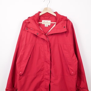 Puede incluir: Chaqueta roja brillante con capucha, cierre de cremallera y botones de presión. La chaqueta tiene dos bolsillos delanteros y un cordón en el dobladillo. Cuelga de una percha de madera.