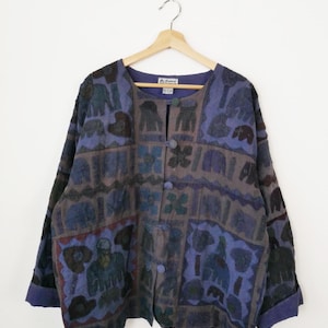 May include: A vintage button-down jacket in blue, grey, and brown. The jacket has a patchwork design with various shapes and patterns, including floral and animal motifs. It has a round neckline and long sleeves with rolled cuffs.
