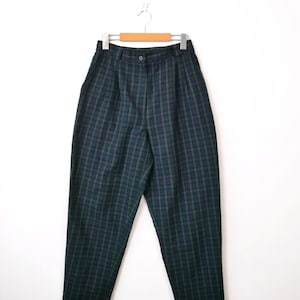 May include: High-waisted trousers featuring a dark green and navy blue plaid pattern. These pants include a front button closure, a waistband with belt loops, and subtle pleats at the waist, hanging from a wooden hanger against a plain background.