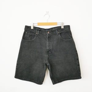 May include: A pair of black denim shorts featuring a classic five-pocket design, a button and zipper fly closure, and belt loops around the waistband. The shorts are displayed on a wooden hanger against a plain white background.