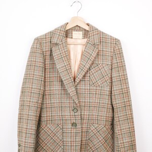 Peut inclure: Un blazer vintage à carreaux marron, orange et vert, suspendu à un cintre en bois. Le blazer présente un col cranté classique, deux poches avant et deux boutons. La doublure intérieure est de couleur pêche clair.