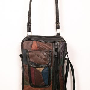 May include: A rectangular crossbody bag constructed from multi-coloured leather patches in shades of brown, black, blue, and burgundy. The bag features a top zip closure, a front flap pocket, a vertical side zip pocket, and a thin black shoulder strap.