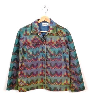 May include: A denim jacket with a colourful geometric pattern in teal, purple, red, and yellow. The jacket has two chest pockets, button closures, and a collar. The label reads "Chico's Design".