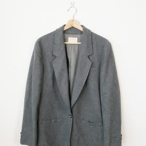 Pode incluir: Um blazer cinza com fecho de um botão, dois bolsos frontais e lapela entalhada. O blazer está pendurado em um cabide de madeira contra um fundo branco. As mangas têm botões nos punhos.