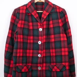 May include: A red and green plaid blazer with three large, round, silver buttons. The blazer has two front pockets and is hanging on a wooden hanger. The plaid pattern is a classic design.