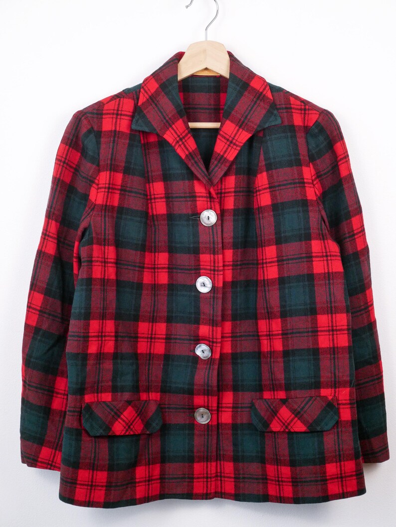 May include: A red and green plaid blazer with three large, round, silver buttons. The blazer has two front pockets and is hanging on a wooden hanger. The plaid pattern is a classic design.
