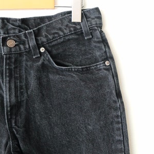 May include: A pair of black denim jeans featuring a high-waisted cut, a metal button closure at the waist, and a classic five-pocket design with silver-toned metal rivets. The fabric has a slightly faded, washed-out texture throughout the material.
