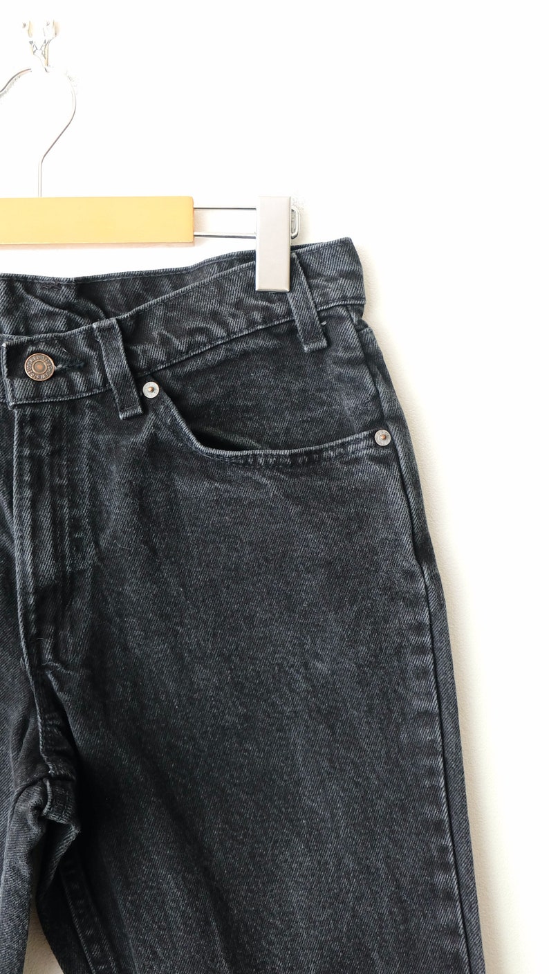 May include: A pair of black denim jeans featuring a high-waisted cut, a metal button closure at the waist, and a classic five-pocket design with silver-toned metal rivets. The fabric has a slightly faded, washed-out texture throughout the material.