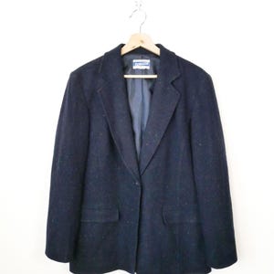 Könnte beinhalten: Ein marineblauer Blazer mit einem Ein-Knopf-Verschluss, zwei Vordertaschen und einem fallenden Revers. Der Blazer hängt an einem Holzbügel vor weißem Hintergrund. Das Etikett im Blazer ist sichtbar.