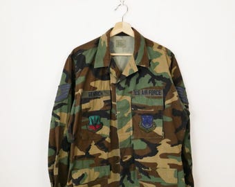 May include: A camouflage military jacket with a name tag that reads "Genrich" and a patch that reads "U.S. Air Force". The jacket has two patches on the front, one with a blue, red, and green design and the other with a blue design.