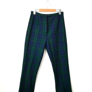 May include: High-waisted trousers featuring a dark green and navy blue plaid tartan pattern. The trousers have a straight-leg cut, a structured waistband with a centre front button closure, and are displayed hanging on a wooden clothes hanger.