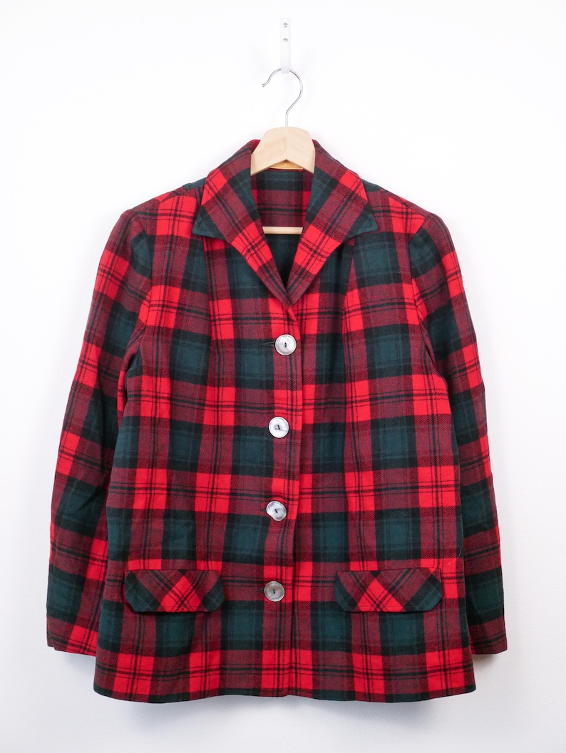 May include: A red and green plaid blazer with three buttons and two front pockets. The jacket is hanging on a wooden hanger against a white background. The plaid pattern is a classic design.
