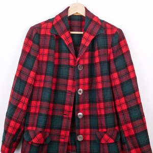 May include: A red, green, and black plaid blazer with three large, round buttons. The blazer has a notched collar and two front pockets. The garment is hanging on a wooden hanger against a white background.