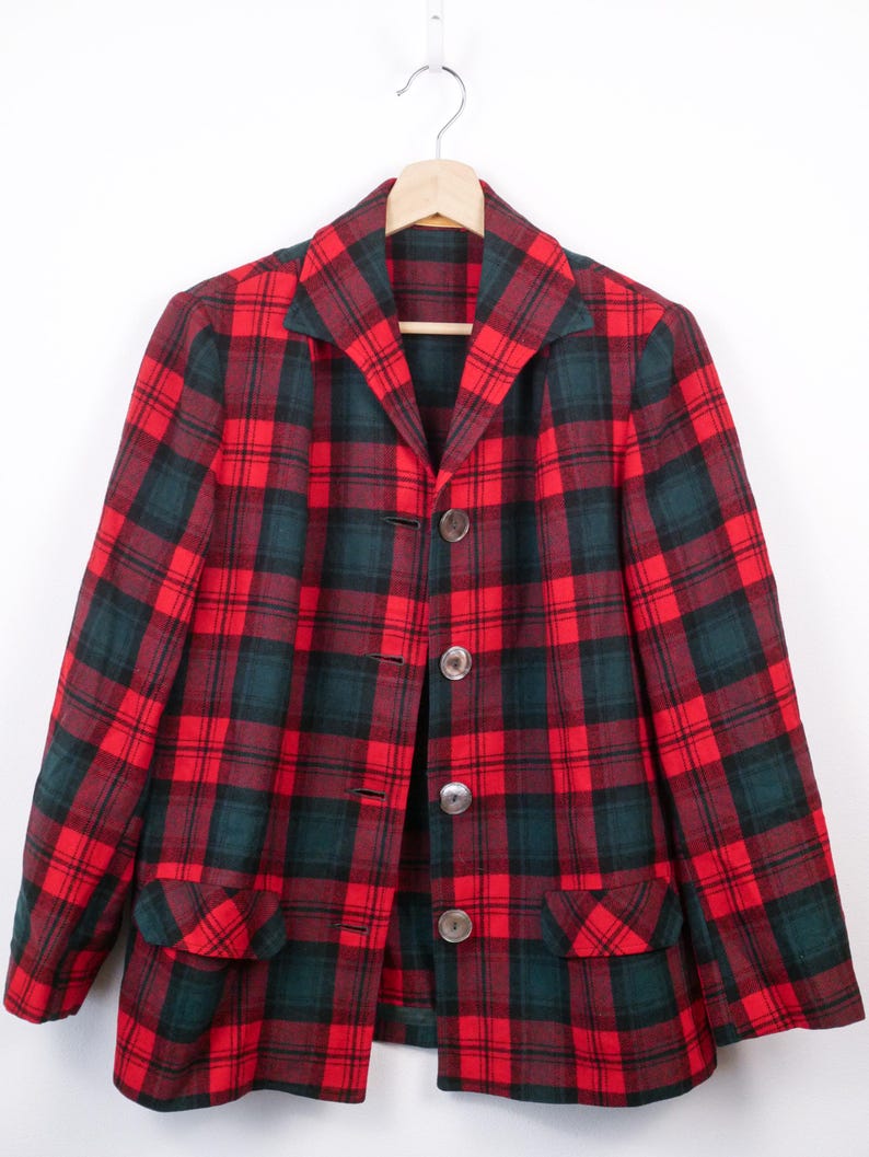May include: A red, green, and black plaid blazer with three large, round buttons. The blazer has a notched collar and two front pockets. The garment is hanging on a wooden hanger against a white background.