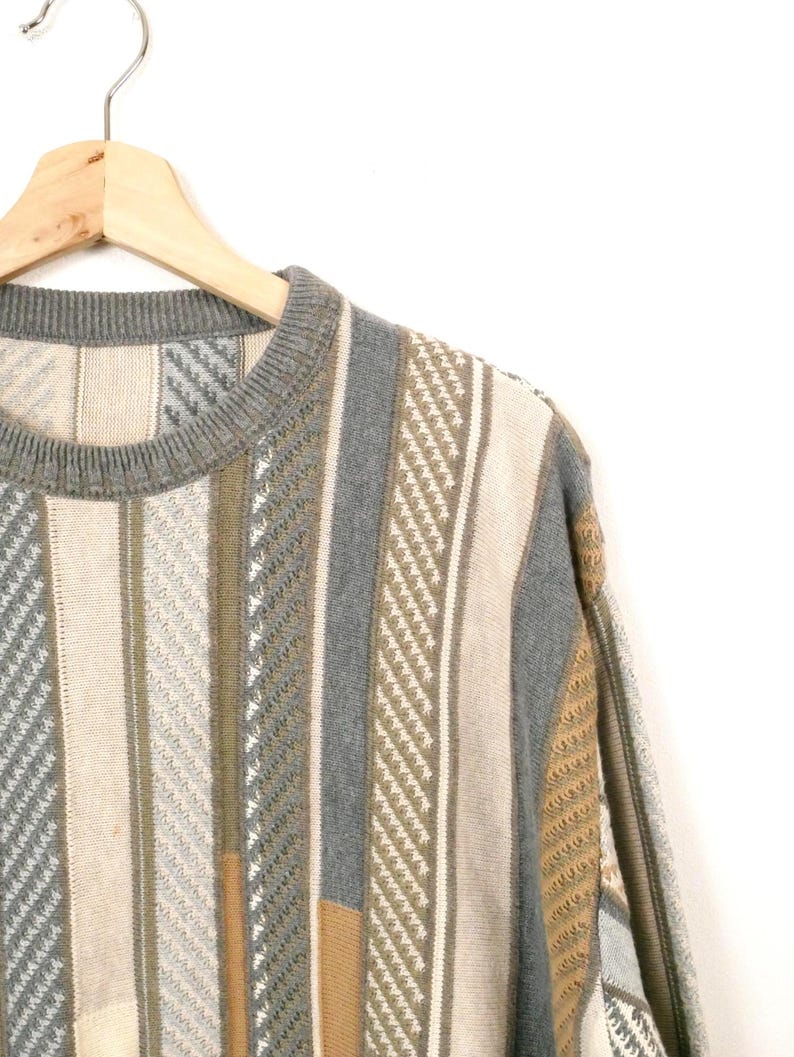 Puede incluir: Un su&eacute;ter vintage con rayas verticales en beige, gris y verde oliva. El su&eacute;ter est&aacute; colgado en una percha de madera sobre un fondo blanco. El patr&oacute;n de punto incluye varias texturas y dise&ntilde;os.
