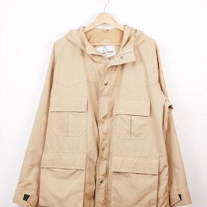 Peut inclure: Veste à capuche de couleur beige avec plusieurs poches avant et une fermeture boutonnée. La veste a un cordon de serrage à la taille et le texte "PACIFIC TRAIL" sur l'étiquette. Suspendue à un cintre en bois.