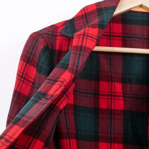 May include: A red and black plaid flannel shirt hanging on a wooden hanger. The shirt's pattern features a classic grid design with alternating red and black squares, and a touch of dark green. The collar and inner lining are visible.