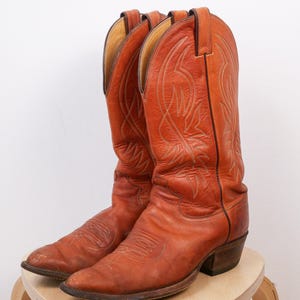Könnte beinhalten: Ein Paar abgenutzte, rotbraune Cowboy-Stiefel mit aufwendigen Stichmustern. Die Stiefel sind aus Leder gefertigt und haben einen klassischen Western-Stil mit spitzen Zehen und gestapelten Absätzen. Sie stehen auf einem kleinen, hellen Holzschemel.