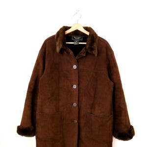 May include: A brown suede coat with a faux fur collar and cuffs. The coat has a button-up front, two front pockets, and is hanging on a wooden hanger. The coat is a medium length.