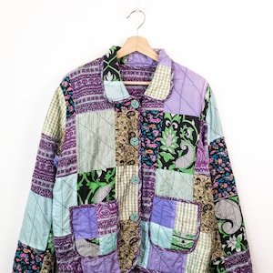 May include: A quilted patchwork jacket featuring a variety of fabric squares in shades of purple, light blue, green, and black. The jacket has a collared neckline, long sleeves, two front patch pockets, and four light green buttons down the front.