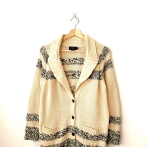 May include: Cream-coloured cardigan sweater with black and grey striped accents. The sweater has a shawl collar, two front pockets, and black buttons. The garment is hanging on a wooden hanger against a white background.