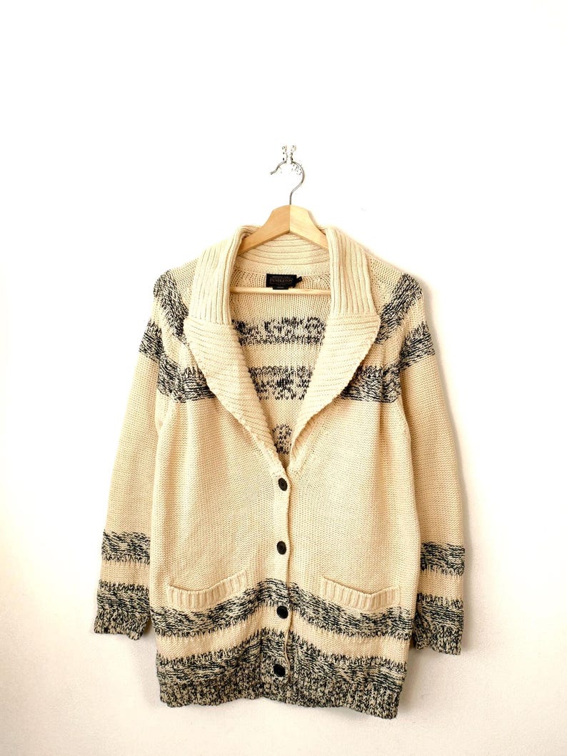 May include: Cream-coloured cardigan sweater with black and grey striped accents. The sweater has a shawl collar, two front pockets, and black buttons. The garment is hanging on a wooden hanger against a white background.