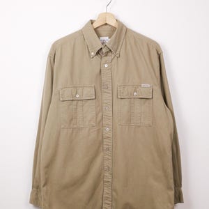 Peut inclure: Une chemise boutonnée beige avec deux poches de poitrine et un petit logo. La chemise à manches longues est suspendue à un cintre en bois sur fond blanc. La chemise a un col classique et des poignets boutonnés.