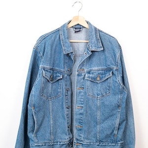 May include: A medium-wash blue denim jacket featuring a classic collar, long sleeves with buttoned cuffs, and two chest pockets with button-down flaps. The jacket has a front button closure with metallic buttons and visible stitching throughout.