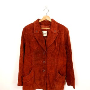 Peut inclure: Veste en daim de couleur rouille avec un col cranté, deux poches avant et des boutons. La veste est suspendue à un cintre en bois sur fond blanc. Une étiquette est visible à l'intérieur de la veste.