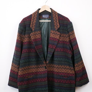 May include: A patterned blazer in shades of burgundy, brown, green, and navy blue. The jacket has a geometric design with horizontal stripes and zigzags. It features a single-button closure and two front pockets. The blazer is displayed on a wooden hanger.
