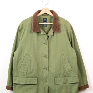 May include: An olive green jacket featuring a brown corduroy collar and matching brown corduroy cuffs. The jacket has a button-front closure, two large flap pockets at the waist, and two vertical slit pockets above them. A label reads Basic Editions.