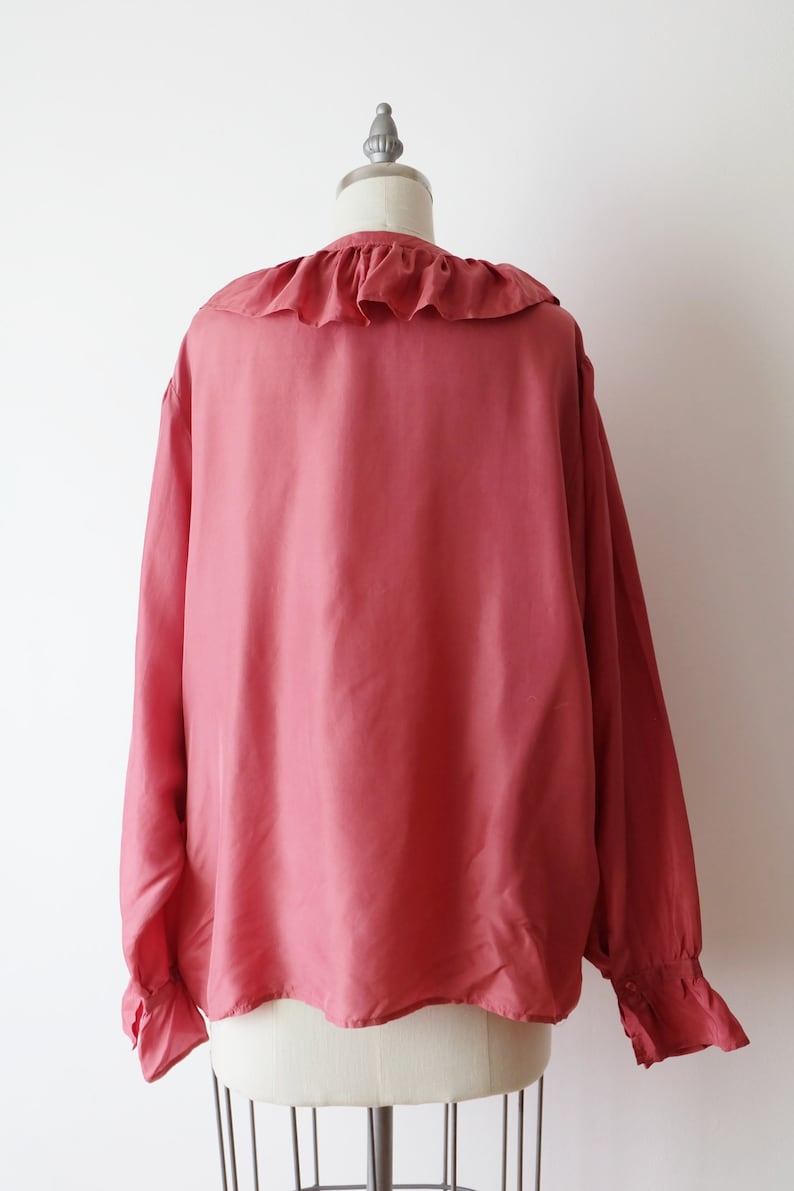 May include: A long-sleeved, salmon-colored blouse with a ruffled collar and cuffs. The blouse is made of a soft, silky fabric.