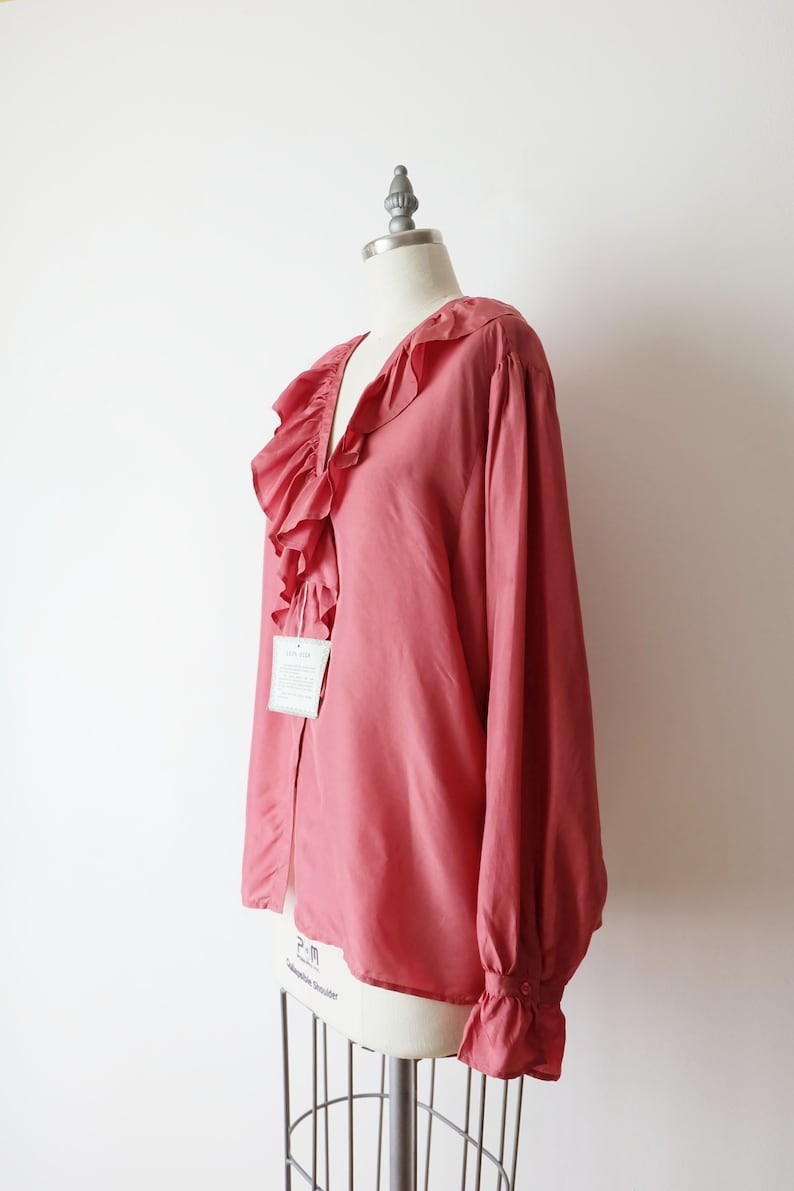 May include: A salmon-colored blouse with a large ruffled collar and long sleeves. The blouse has a button closure and a tag that says "Silk 100%".