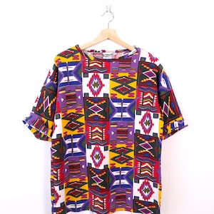 May include: A short-sleeved, patterned shirt with a vibrant geometric design. The shirt features a mix of colors including yellow, purple, red, and white. The design is made up of various shapes and patterns, creating a bold and eye-catching look. The shirt is hanging on a wooden hanger.