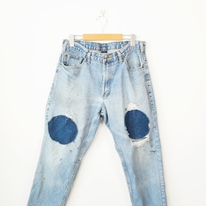 May include: Light blue denim jeans with two large, dark blue patches on the knees. The jeans show signs of wear and tear, with distressed areas and a faded appearance. They are hanging on a wooden hanger against a white background.