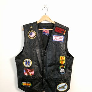 Peut inclure: Gilet en cuir noir orné de nombreux écussons, dont un drapeau américain, des insignes militaires et le texte « J'aime mon pays mais je crains mon gouvernement ». Le gilet a une fermeture boutonnée et deux poches avant.