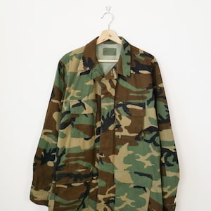 Pode incluir: Casaco com padrão de camuflagem com fecho de botões, dois bolsos no peito e mangas compridas. O casaco está pendurado num cabide de madeira sobre um fundo branco. O design de camuflagem apresenta tons de verde, castanho e preto.