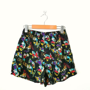 Op de afbeelding: Zwarte korte broek met een kleurrijk patroon met stripfiguren en het woord "FORE!". De korte broek hangt aan een hanger tegen een witte achtergrond. De elastische tailleband is zichtbaar.