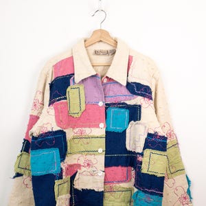 May include: A long-sleeve button-up jacket featuring a patchwork design with cream, navy blue, pink, light green, and turquoise fabric squares. The garment includes visible raw edges, decorative pink floral embroidery, and a cream-coloured collar.