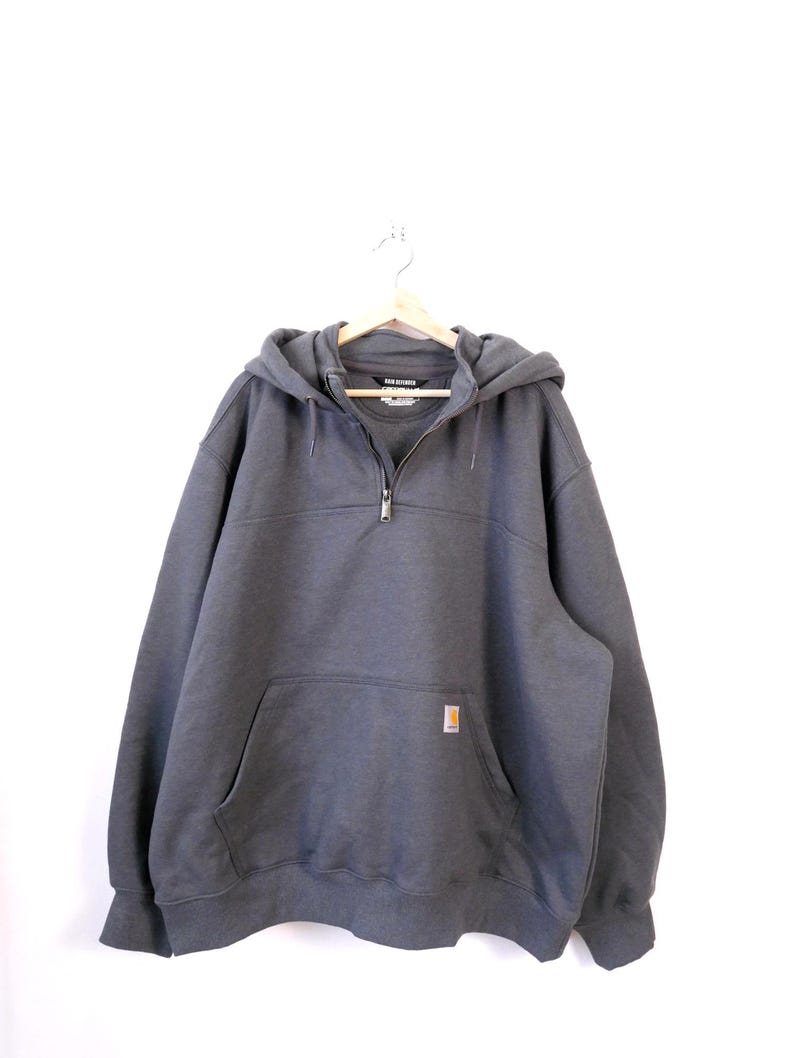May include: A charcoal gray hooded sweatshirt with a half-zip closure and a front kangaroo pocket. The sweatshirt features a small, rectangular logo patch on the pocket and is hanging on a wooden hanger.