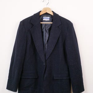 May include: A dark navy blue blazer with a subtle speckled texture, featuring a single-button closure, notched lapels, and two front flap pockets. The interior lining is dark blue and includes a Pendleton brand label at the neckline.
