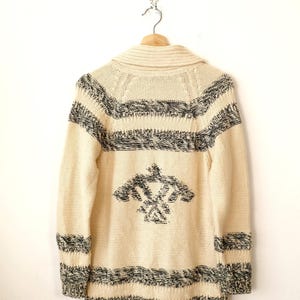 May include: Cream-coloured knitted cardigan with black and white geometric patterns. The long-sleeved sweater features a shawl collar and a decorative design on the back. The cardigan is hanging on a wooden hanger.