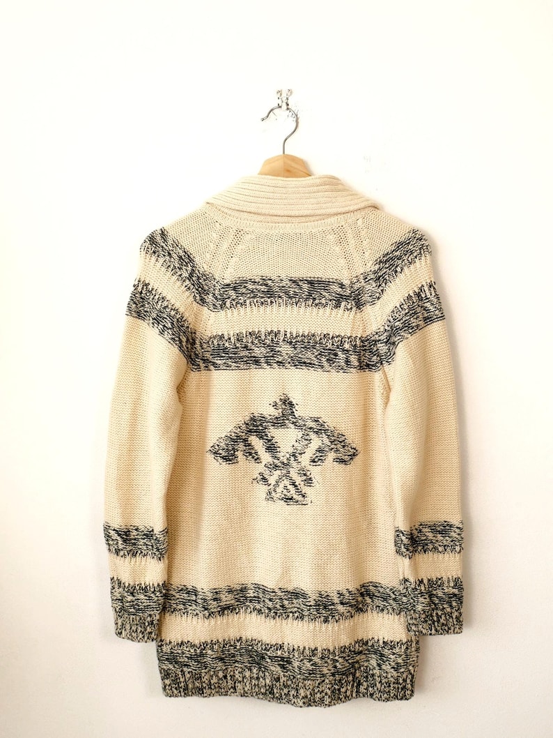 May include: Cream-coloured knitted cardigan with black and white geometric patterns. The long-sleeved sweater features a shawl collar and a decorative design on the back. The cardigan is hanging on a wooden hanger.