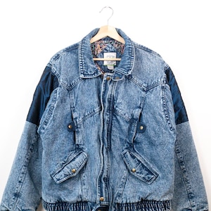 May include: A blue acid-wash denim jacket featuring dark blue satin panels on the shoulders, a fold-over collar, and two front flap pockets with brass-toned buttons. The jacket has a ribbed elastic waistband and cuffs, with a patterned inner lining.