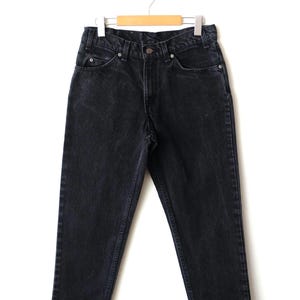 May include: A pair of black denim jeans featuring a high-waisted design, a button and zipper fly closure, five pockets, and belt loops. The jeans are constructed with a straight-leg cut and are displayed hanging from a wooden clothes hanger.