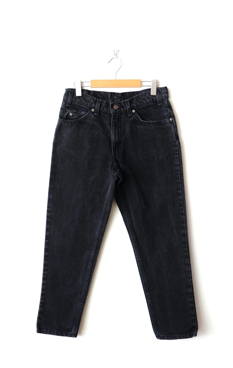 May include: A pair of black denim jeans featuring a high-waisted design, a button and zipper fly closure, five pockets, and belt loops. The jeans are constructed with a straight-leg cut and are displayed hanging from a wooden clothes hanger.