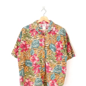 Può includere: Una camicia a maniche corte con bottoni e un vivace motivo floreale. La camicia presenta grandi fiori di ibisco rosa, foglie verde acqua e verdi e uno sfondo beige. La camicia è appesa a una gruccia di legno su sfondo bianco.