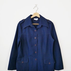 May include: A dark navy blue jacket with a button-up front, two front pockets, and shoulder epaulets. The jacket is suspended on a wooden hanger. The buttons and pocket details are a darker shade of blue. The background is white.